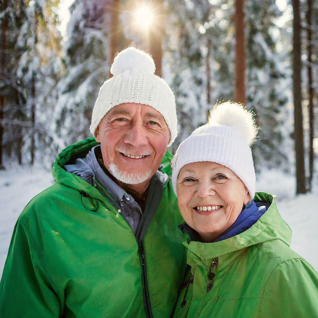 Vanha pariskunta hymyilee vihreissä takeissa ja valkoiset myssyt päässään. Taustalla talvinen metsä.