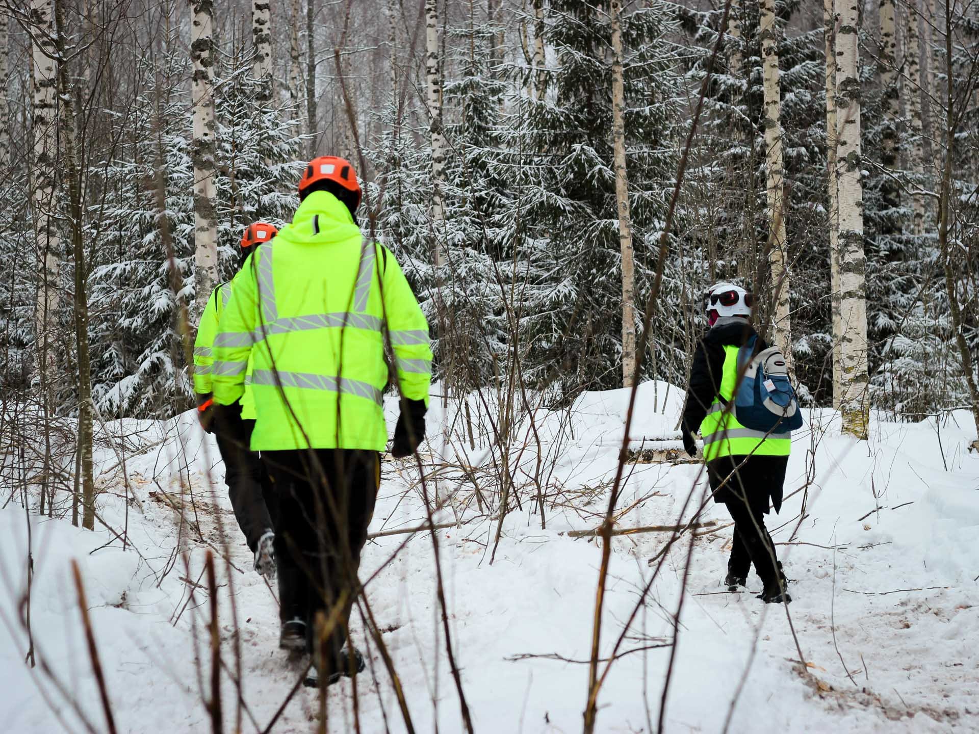 Kolme kirkkaisiin vaatteisiin sonnustautunutta henkilöä kävelee metsässä ajouria pitkin.