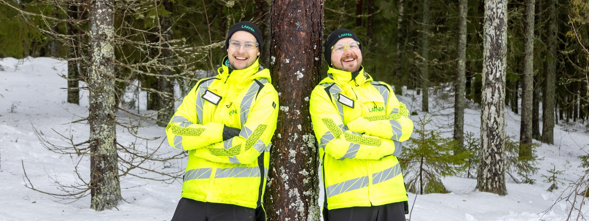 Kaksi metsäasiantuntijaa nojaavat puuhun ja poseeraavat hymyillen.