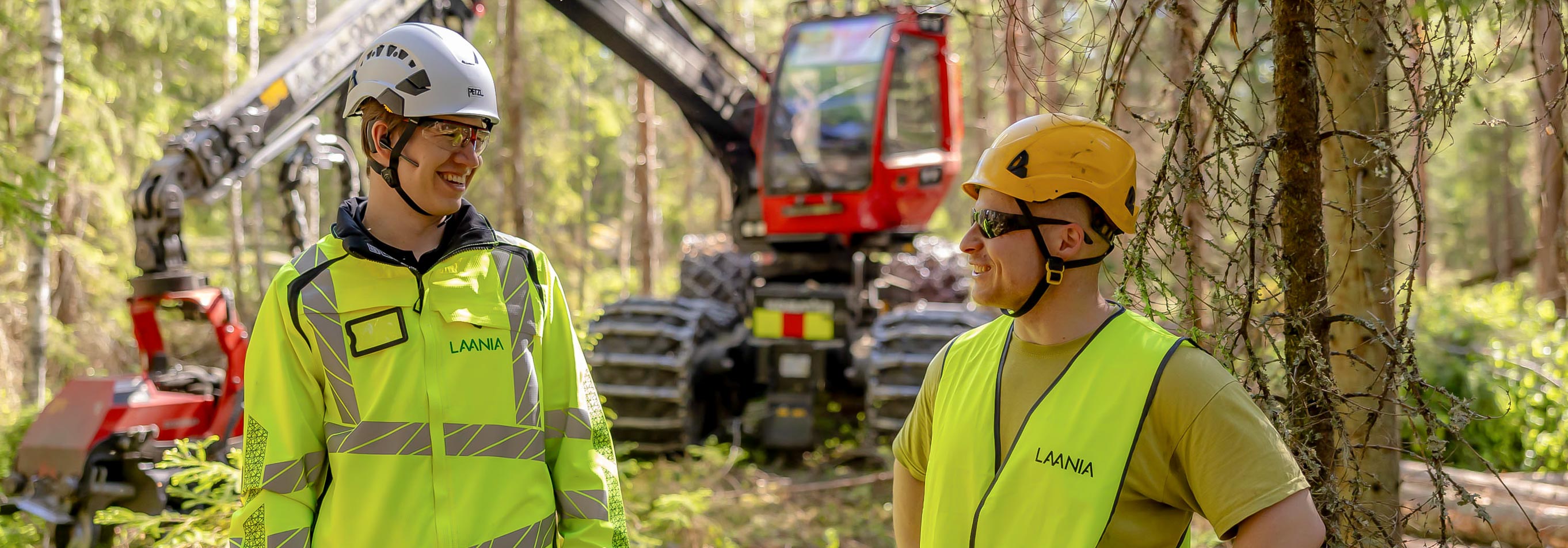 Kaksi henkilöä katsovat toisiaan hymyillen metsässä, taustalla näkyy metsäkone.